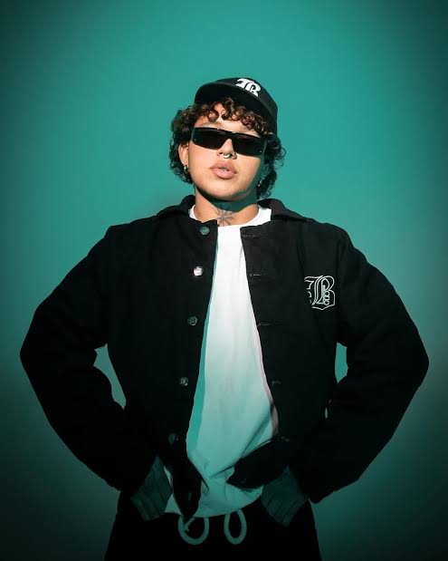 Beéle, cantante barranquillero, posa con estilo urbano luciendo una chaqueta negra y gafas de sol sobre un fondo verde.