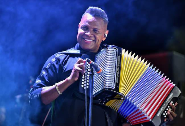 Omar Geles sonríe mientras toca su acordeón en un concierto, con fondo iluminado y ambiente musical vallenato.